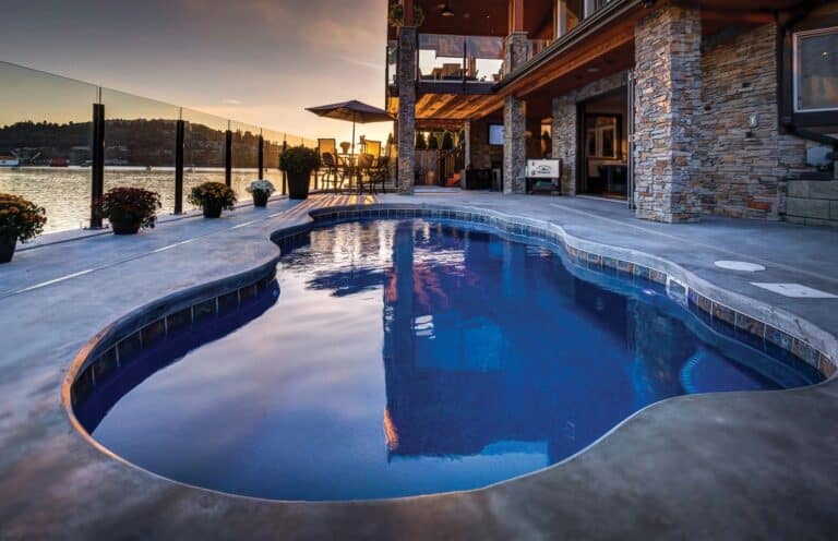 Freeform Aruba fiberglass pool with dark blue finish and stone coping at sunset beside a stone-faced home.